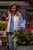 Woman wearing blue shirt, white fuzzy vest, and jeans standing among flowers, holding tan bag, smiling in front of flower shop.