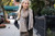 Woman wearing Malabrigo Verano scarf, black jacket, and dress standing on sidewalk with hands in pockets, showcasing beige and brown pima cotton accessory.
