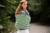 Woman holding green and white striped tote bag, Malabrigo Verano Pima Cotton Collection, outdoors on a path surrounded by trees and foliage.