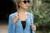 Woman wearing blue cardigan, Malabrigo Verano Pima Cotton, with black top, sunglasses, and braid, outdoors near greenery and tunnel entrance.