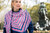 Woman wearing Malabrigo Verano scarf in pink, blue and white stripes, paired with blue outfit standing in front of statue and trees.