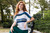 Woman with long red hair wearing white short-sleeved top with blue and green stripes and green skirt sitting on a black chair outdoors.