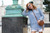 Woman with red hair wearing blue sweater, white shorts, and brown purse standing beside green metal monument base outdoors in summer.