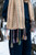 Multicolored tassels hang from a beige knitted scarf, worn with dark blue clothing, in a snowy outdoor setting with trees.