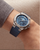 Wrist shot of Baume & Mercier Riviera Tideograph 10761 — dodecagonal polished steel case reflecting light across facets, vivid blue dial and tide disc accentuating nautical sophistication.