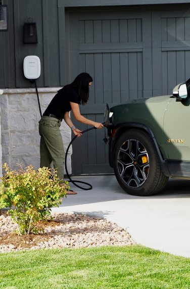 Woman charging electric vehicle in driveway with EV charger mounted to wall.