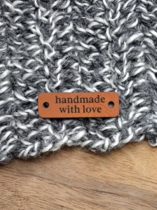 Single custom chestnut faux leather tag engraved with "handmade with love", positioned flat on a textured grey and white knitted fabric with an oak wood background