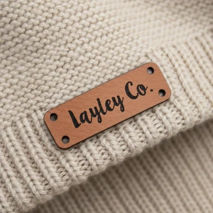Single custom chestnut faux leather tag engraved with customer design, positioned flat on a textured beige knitted fabric