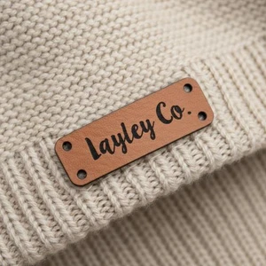 Custom Faux Leather Labels with Your Own Design | 0.5" x 1.5" Corner Holes Sew-On Tags (set of 25), tan faux leather label with black script lettering and four corner holes sewn onto a cream knitted garment