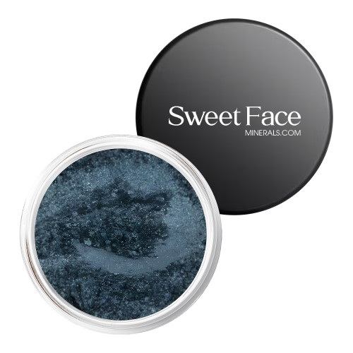 An open jar of Sweet Face Minerals Smolder loose mineral eyeshadow showcasing a deep slate blue shade with a cool metallic shimmer, paired with the branded black lid on a clean white background.