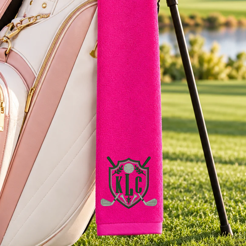 pink golf towel with a black KGS shield monogram, crossed golf clubs, and silver golf ball hanging from a golf bag.