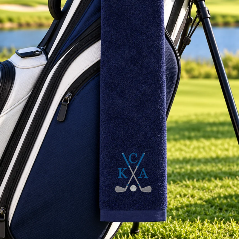Navy golf towel embroidered with three blue initials around crossed golf clubs and a silver golf ball, hanging on a golf bag.