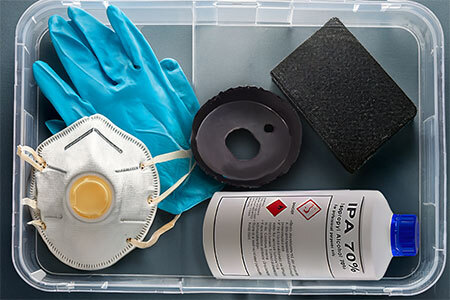 Flat-lay of Still Air Box building materials including a clear plastic tote, hole saw, sandpaper, isopropyl alcohol spray, nitrile gloves, and face mask