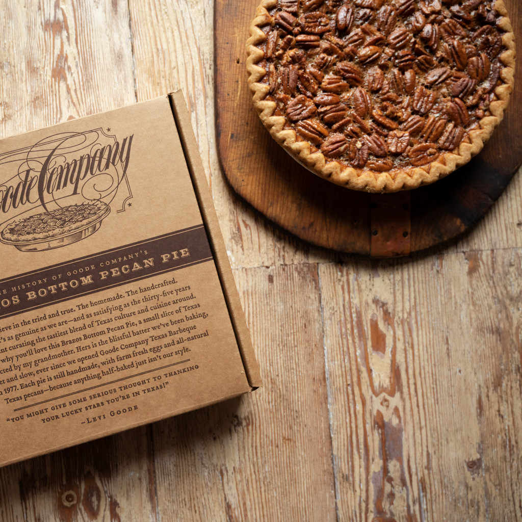 A pecan pie on a wooden table with a carboard box on the side of it that shows the Goode Company logo on the front with a description of the pie.