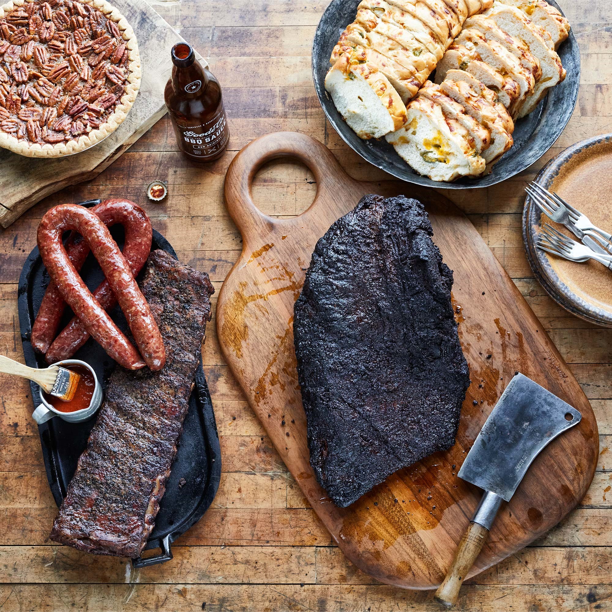 Rustic table with a slab of brisket on a cutting board along with a plated ribs and sausage. A pecan pie and sliced loaf of jalapeno cheese bread are off to the side.