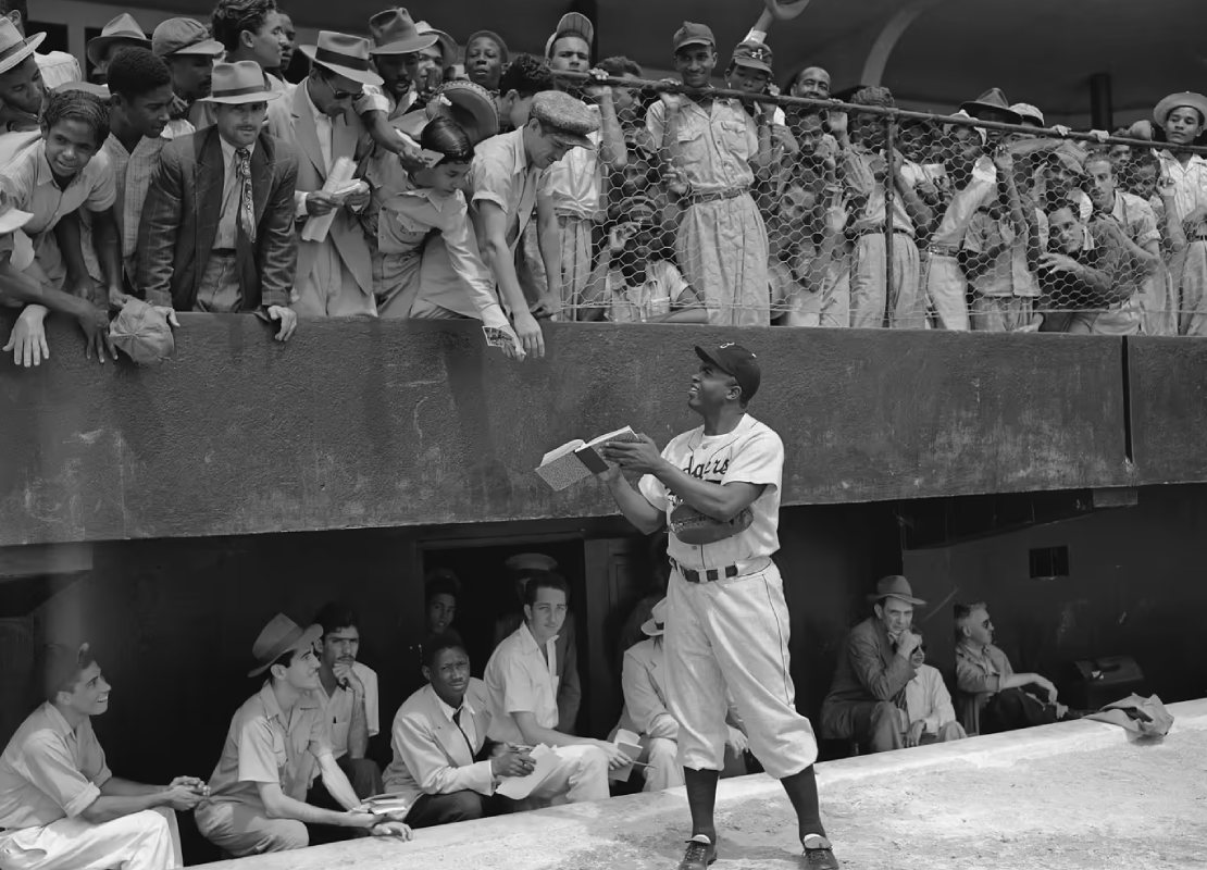 Black and white image of Jackie Robinson signing autographs for a crowd of people during a baseball game