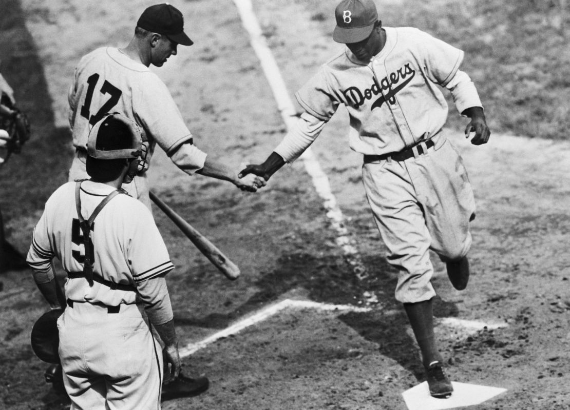 Black and white image of Jackie Robinson shaking hands with fellow teammate as he runs across home base during a Baseball game