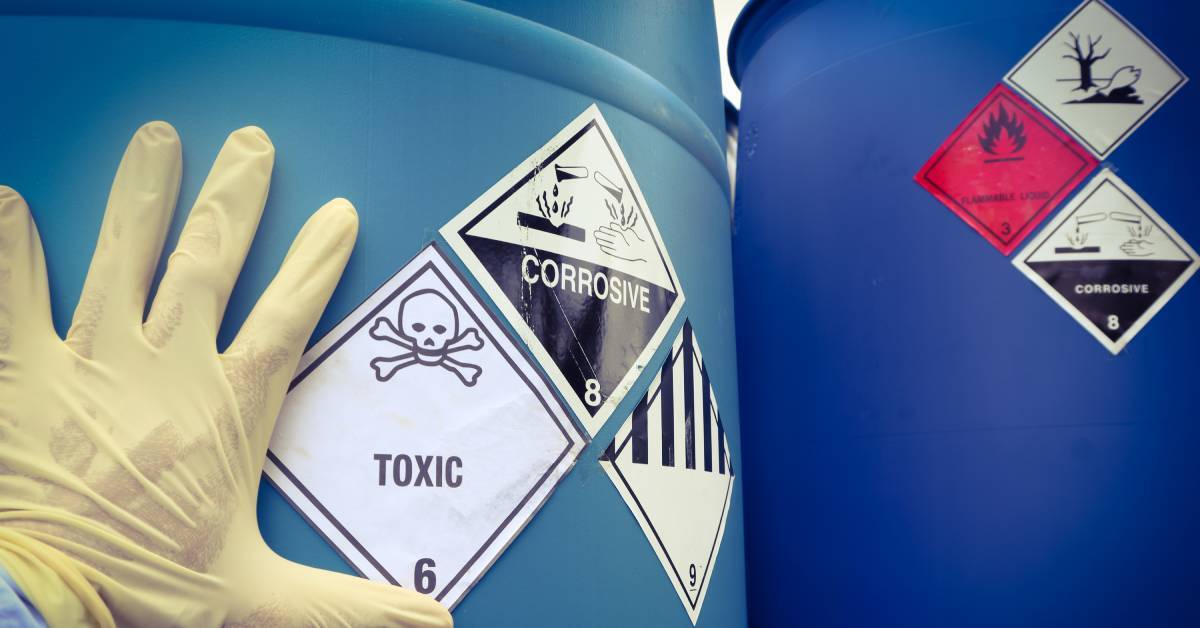 Raised arm wearing protection gloves, touching a large blue container with stickers displaying “toxic” and “corrosive”