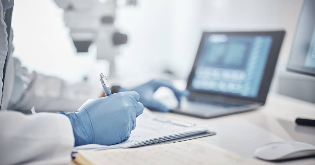 A person in a white coat and blue gloves holds a pen over some documents. They are also working on a laptop.