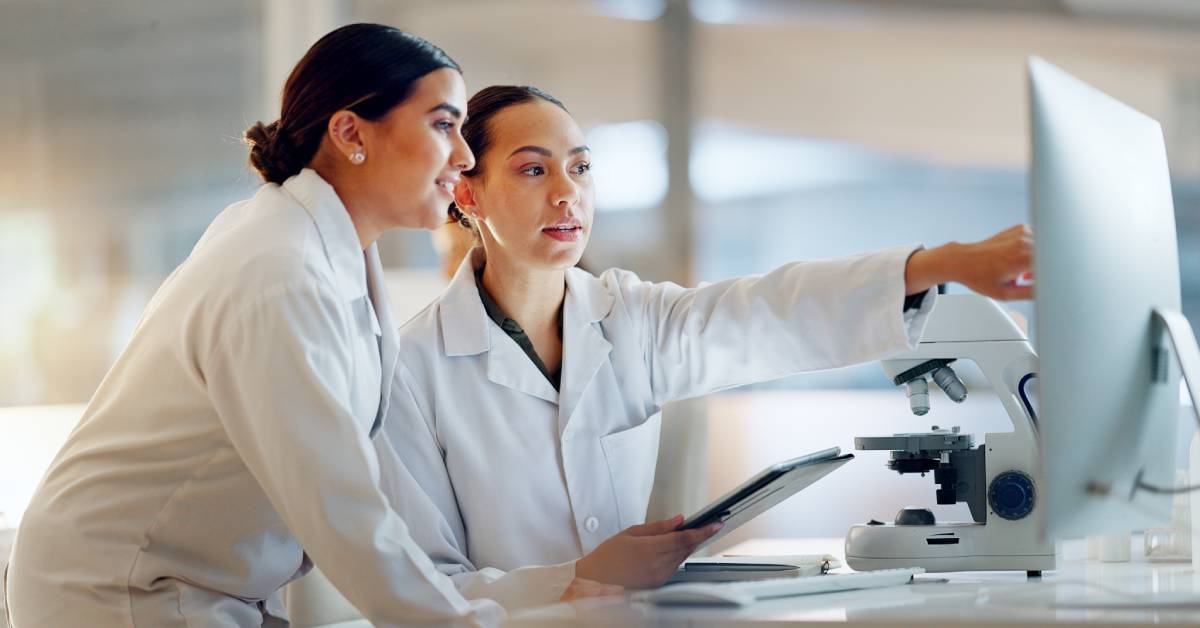 Two women in white lab coats are looking at a computer screen. There is a microscope next to the computer monitor.