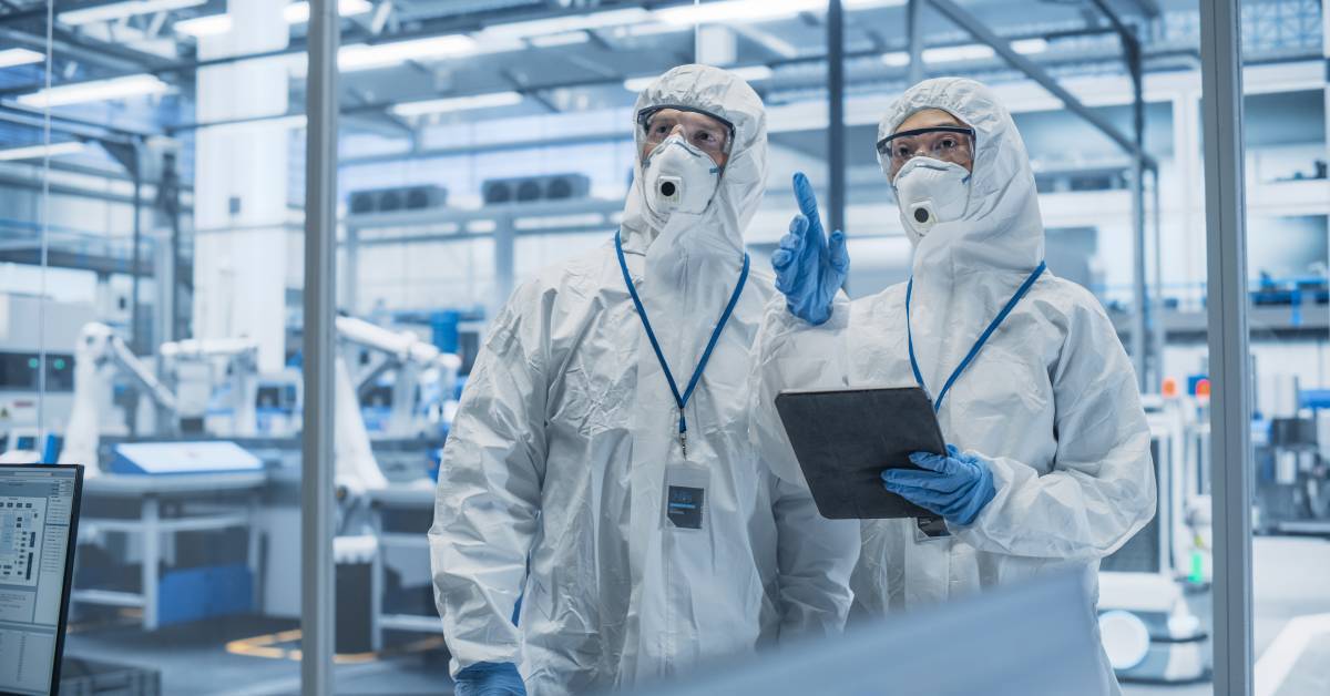 Two scientists standing inside a large modern laboratory wearing white protection suits, glasses, face masks, and gloves.