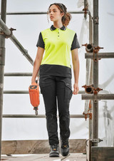 A woman wearing bright yellow and black cargo pants, standing on scaffolding with a power tool, showcasing the cargo pant design.