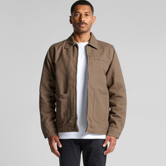 A man wearing a brown canvas heavy jacket with two front pockets and a zippered closure, standing against a grey background.