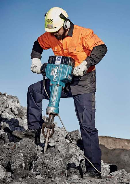 A man in safety gear operates a jackhammer on a rocky surface, wearing an orange shirt and navy blue cargo pants.