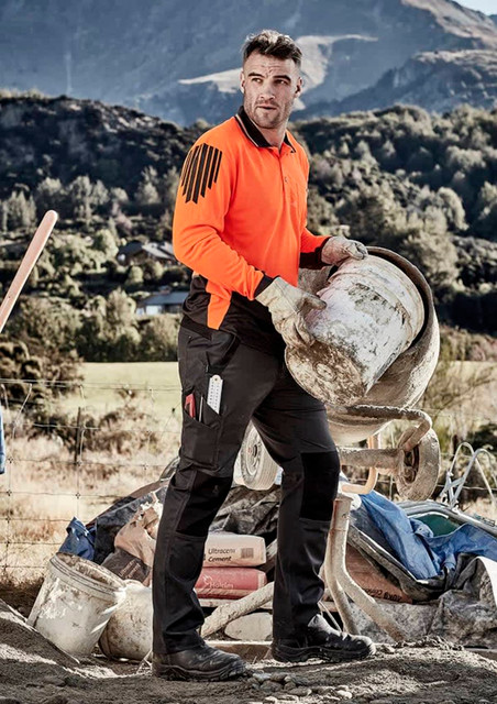 A worker in an orange high-visibility shirt and black tough pants, holding a bucket on a job site with mountains in the background.