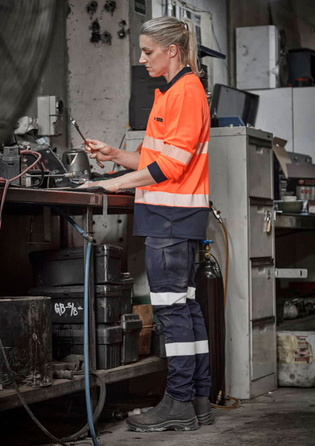 A woman wearing high-visibility orange and navy cargo pants with cuffs, working at a bench in a workshop.