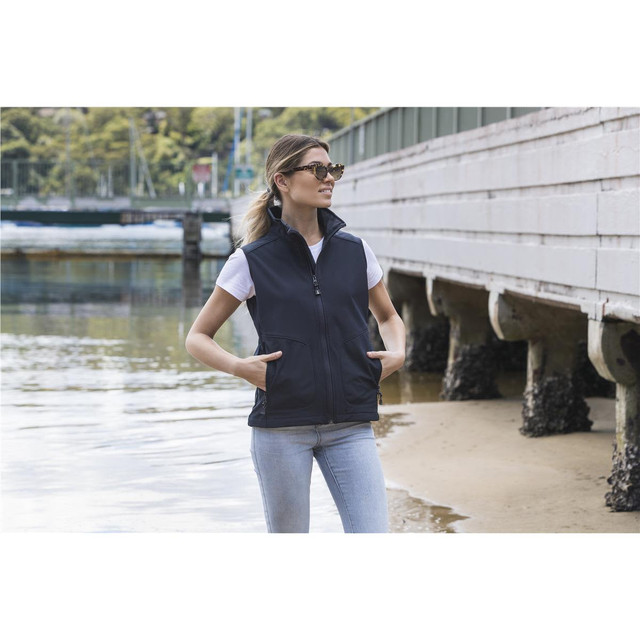 A woman wearing a black softshell vest over a white T-shirt, standing near the water with hands in pockets.