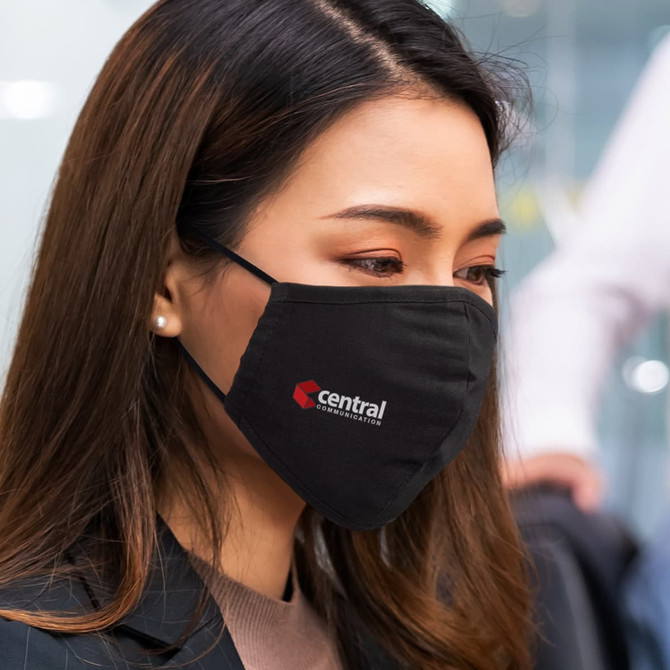 A black face mask featuring a logo, worn by a woman with long hair, in a professional setting.