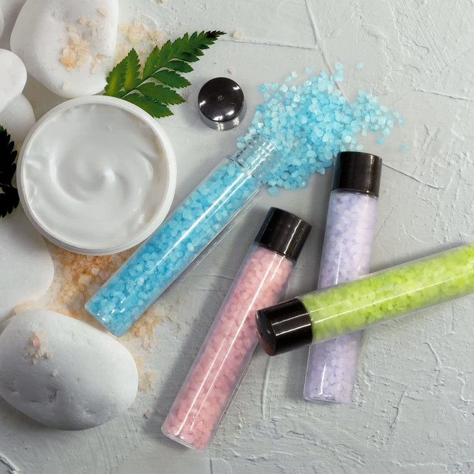 A set of bath salts in clear tubes featuring blue, pink, green, and lavender colours, alongside a white cream in a jar and pebbles.