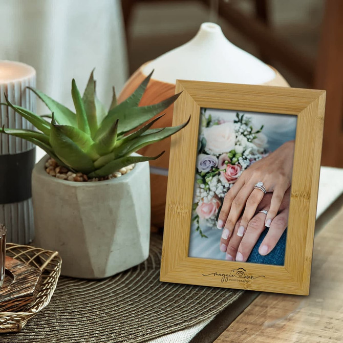 A NATURA wooden photo frame in a light natural finish displays a photo of hands with a ring and flowers, surrounded by decor.