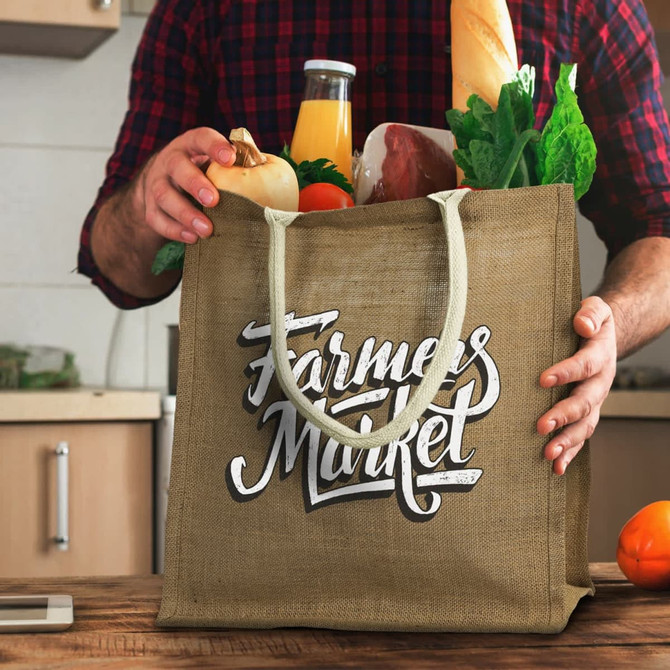 A jute tote bag in a tan colour filled with groceries, including bread, vegetables, and juice, featuring a logo.