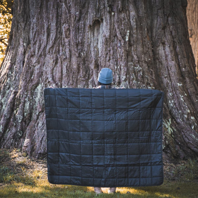 A black puffer blanket is held up by a person in front of a large tree. The scene is outdoors.