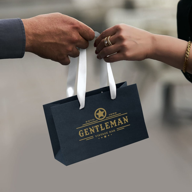 A small black paper bag with a white ribbon handle, featuring a logo in gold.