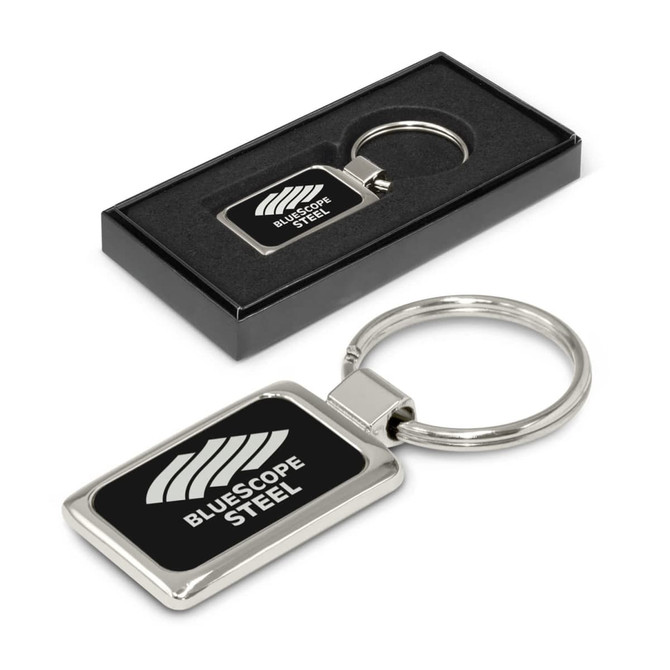 A metal key ring with a rectangular shape featuring a logo. The key ring is presented in a black box.