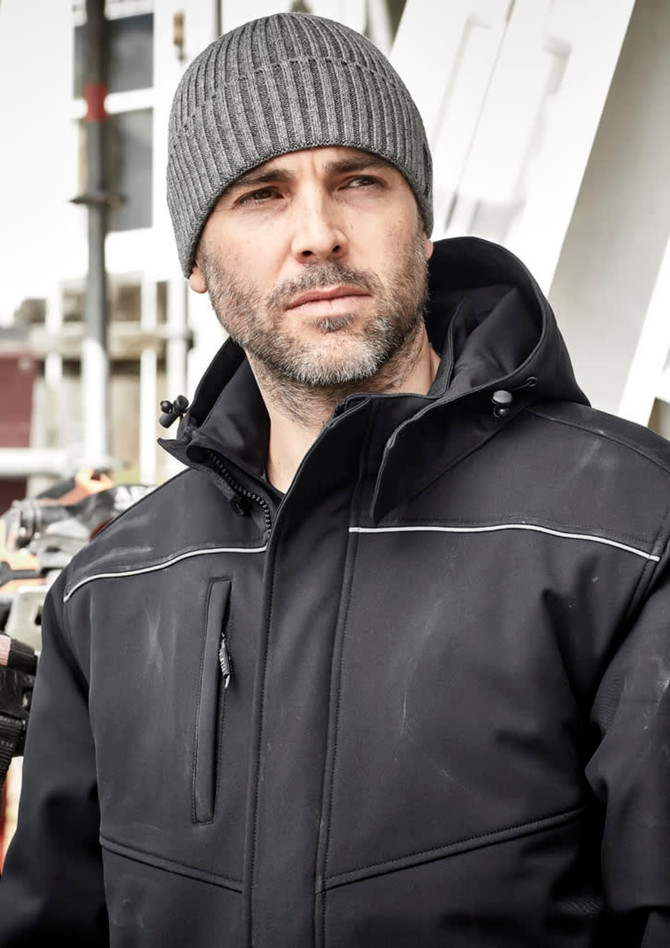 A man in a black softshell jacket and grey beanie, standing in an outdoor setting with construction in the background.