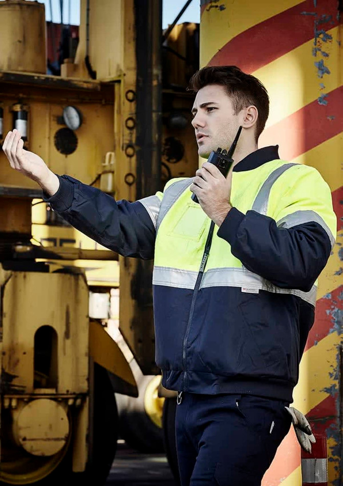 A man in a hi-vis yellow and navy quilted jacket uses a walkie-talkie against an industrial background.
