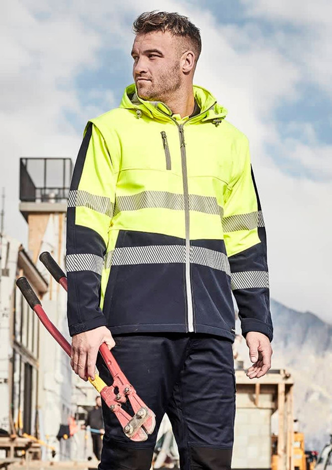 A man in a high-visibility yellow and navy softshell jacket holds red bolt cutters, set against a construction backdrop.