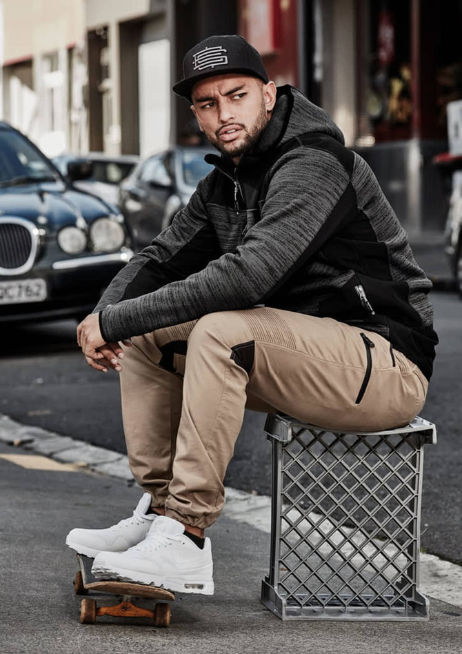 A man sits on a crate wearing cuffed beige stretch pants and a black hoodie. He has white sneakers and a cap.