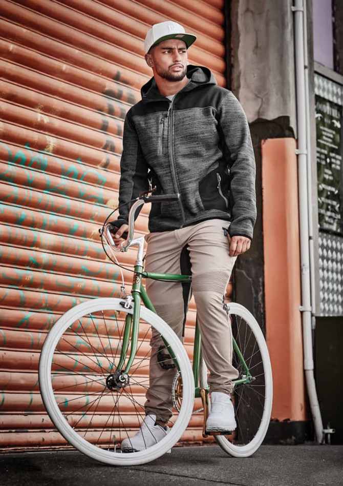 A man wearing cuffed beige stretch pants and a grey jacket stands beside a green bicycle against a textured orange backdrop.