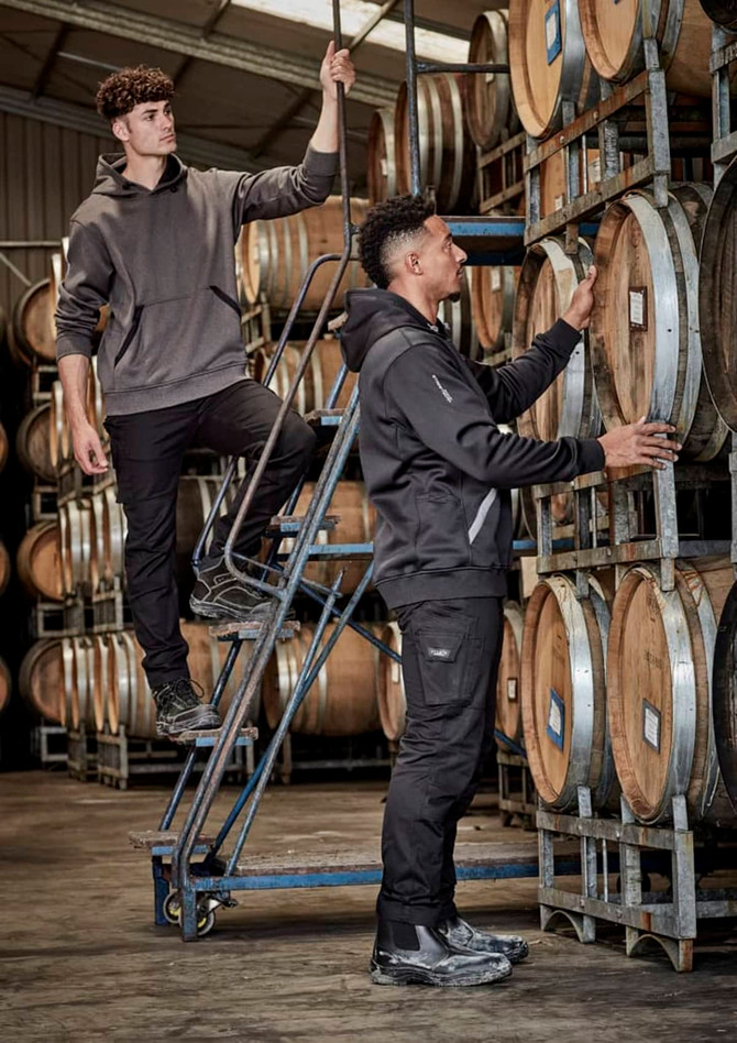 Two men in rugged cooling stretch pants, one standing on a ladder and the other inspecting wooden barrels, in a warehouse setting.