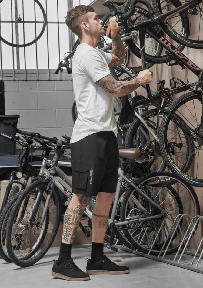 A man stands in a garage adjusting a bike, wearing black Men's Streetworx board shorts and a white t-shirt.