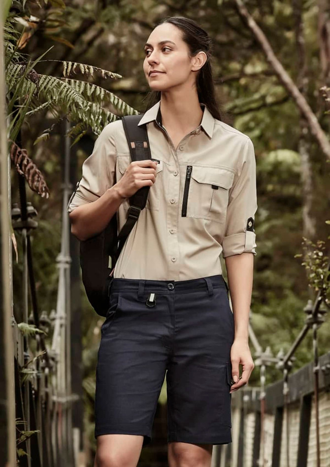 A woman in a light beige shirt and navy shorts stands outdoors, with a backpack over her shoulder. The shorts feature a logo.