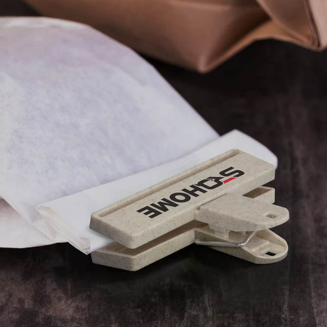 A light grey snack bag clip rests on a dark surface, holding a piece of white paper. It features a logo.