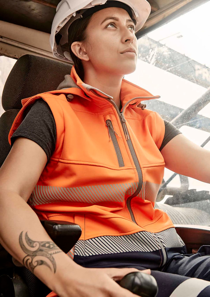 An orange softshell jacket with reflective detailing, worn by a woman, seated in a vehicle with a hard hat.