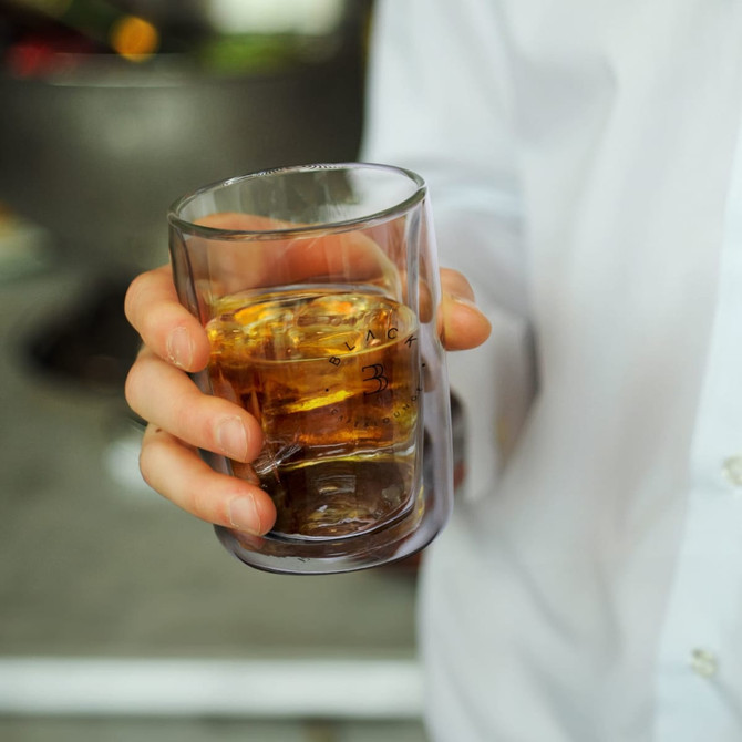 A drink bottle made of double wall glass, featuring a clear design with a brown liquid inside, held in a hand.