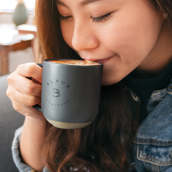 A ceramic mug in black with a textured finish, featuring a logo, held by a person enjoying a beverage.
